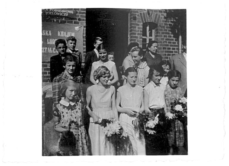 Zdjęcie przedstawia grupę uczniów i nauczyciel na tle wejścia do starej szkoły w Wielowiczu
